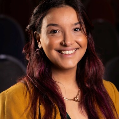 Headshot of Sofi Berenger, long dark hair, smiling and wearing a mustard colour jacket