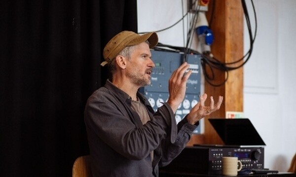 Head shot of Ben Duke wearing a cap behind him a laptop and theatre rehearsal space