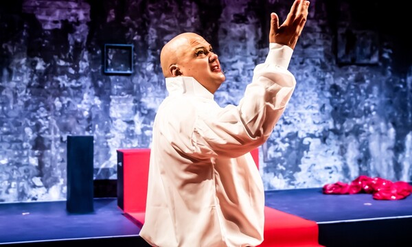 A glittery stage background with red carpet, in the foreground a brown skinned man in a white shirt holds up his hand to the ceiling