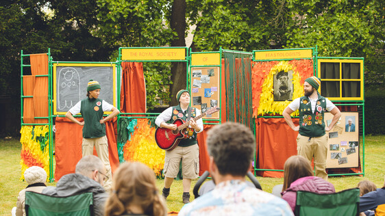 3 performers, one with a guitar standing in front of people sat in camping chairs