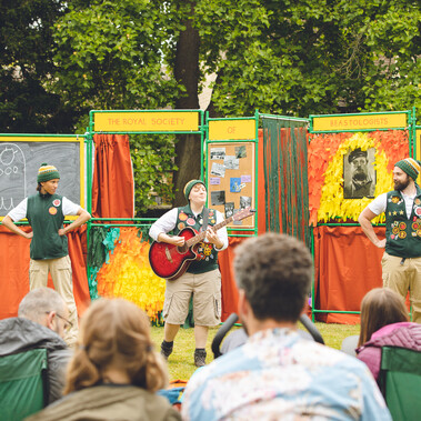 3 performers, one with a guitar standing in front of people sat in camping chairs