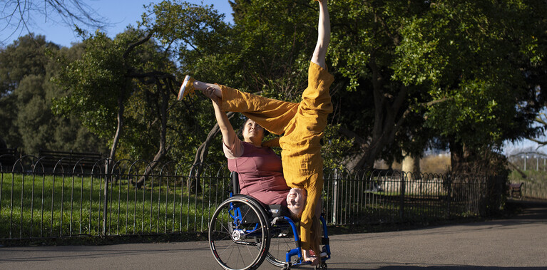 In the centre of the images are 2 performers. Laura, a curvy brown skinned Filipino female wheelchair user with an above right knee amputation is wearing a plum coloured dress. She sat in a manual wheelchair. Danielle, a white woman with ginger hair in ponytails, is wearing an ochre jumpsuit. She is upside down in a one arm handstand position with legs split creating a Y shape with her body. She is supported by Laura who holds one of Danielle's ankles. They look out towards the sunlight smiling. They are on a grey sunlit asphalt path. Behind them are trees and shrubbery and a bright blue sky.