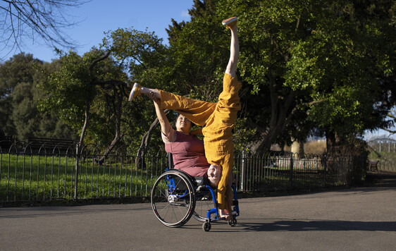 In the centre of the images are 2 performers. Laura, a curvy brown skinned Filipino female wheelchair user with an above right knee amputation is wearing a plum coloured dress. She sat in a manual wheelchair. Danielle, a white woman with ginger hair in ponytails, is wearing an ochre jumpsuit. She is upside down in a one arm handstand position with legs split creating a Y shape with her body. She is supported by Laura who holds one of Danielle's ankles. They look out towards the sunlight smiling. They are on a grey sunlit asphalt path. Behind them are trees and shrubbery and a bright blue sky.